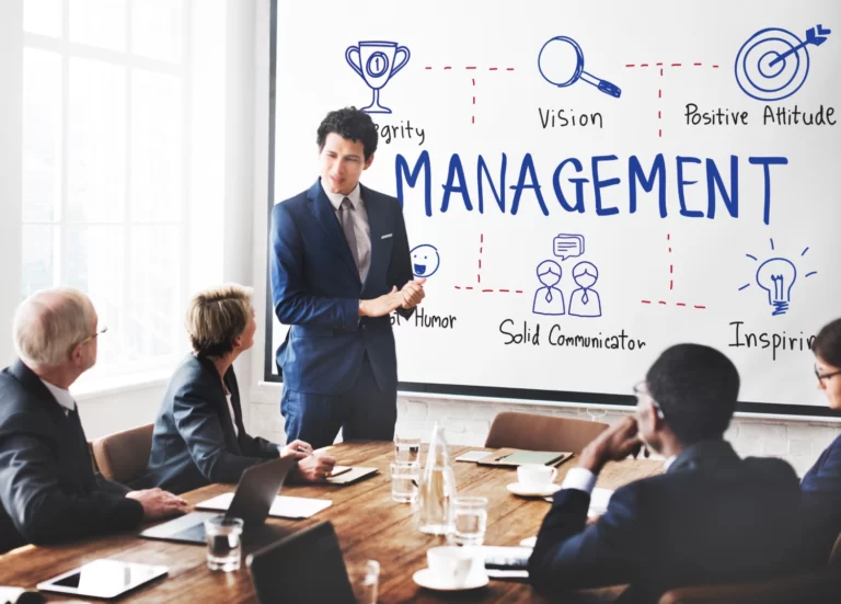 Une personne qui donne une formation en management à un groupe de personnes. Il se trouve devant un tableau blanc avec les valeurs du management inscrites dessus.